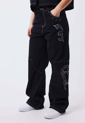 Persona che indossa jeans neri a gamba larga con disegni ricamati bianchi sulla coscia e sul ginocchio, abbinati a scarpe da ginnastica bianche.