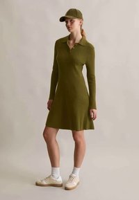 Ženska haljina dugih rukava od rebrastog materijala u maslinasto zelenoj boji s polo ovratnikom, lepršavim rubom, u kombinaciji s odgovarajućom kapom i bijelim tenisicama s bež detaljima.