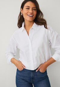 Chemise blanche en coton avec un col classique, des manches longues et une coupe décontractée, rentrée dans un jean en denim bleu foncé.