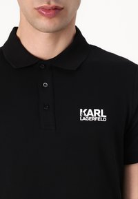 Čierna polokošeľa s klasickým golierom, trojradovým zapínaním na gombíky a bielym logom "KARL LAGERFELD" na ľavej strane hrudníka.