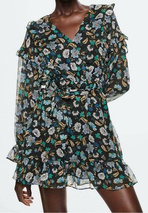 Robe longue à manches longues noire à motif floral bleu, blanc et vert, avec épaules et ourlet volantés, et ceinture nouée à la taille portée par une personne debout.