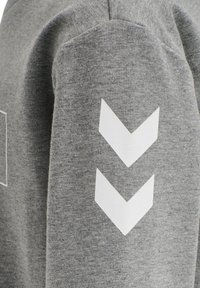 Tessuto grigio con due frecce a forma di chevron bianche che puntano verso il basso stampate sulla manica di un indumento, mostrando dettagli di texture e cucitura.