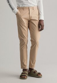 Pantalon en coton beige à coupe slim, avec fermeture à boutons sur le devant et poches latérales, assorti à des sandales vert olive.
