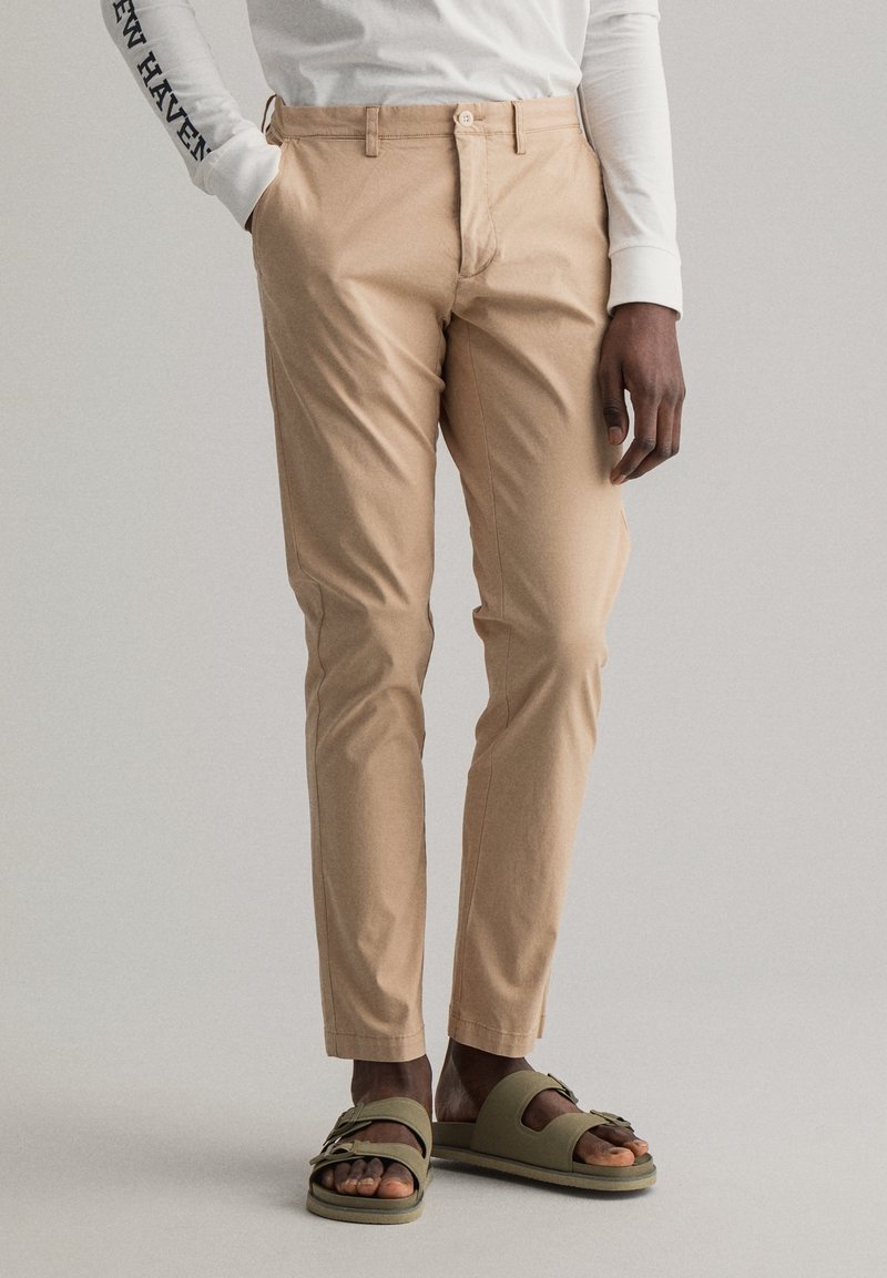 Pantalon en coton beige à coupe slim, avec fermeture à boutons sur le devant et poches latérales, assorti à des sandales vert olive.