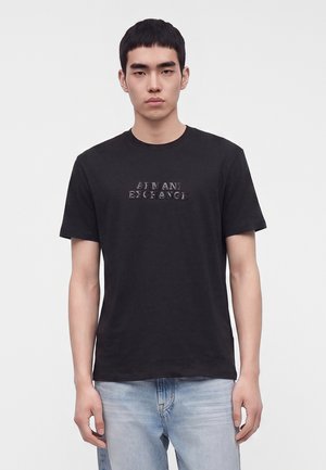 Jeune homme aux cheveux courts noirs portant un t-shirt noir Armani Exchange et un jean bleu clair, se tenant devant un fond clair uni.