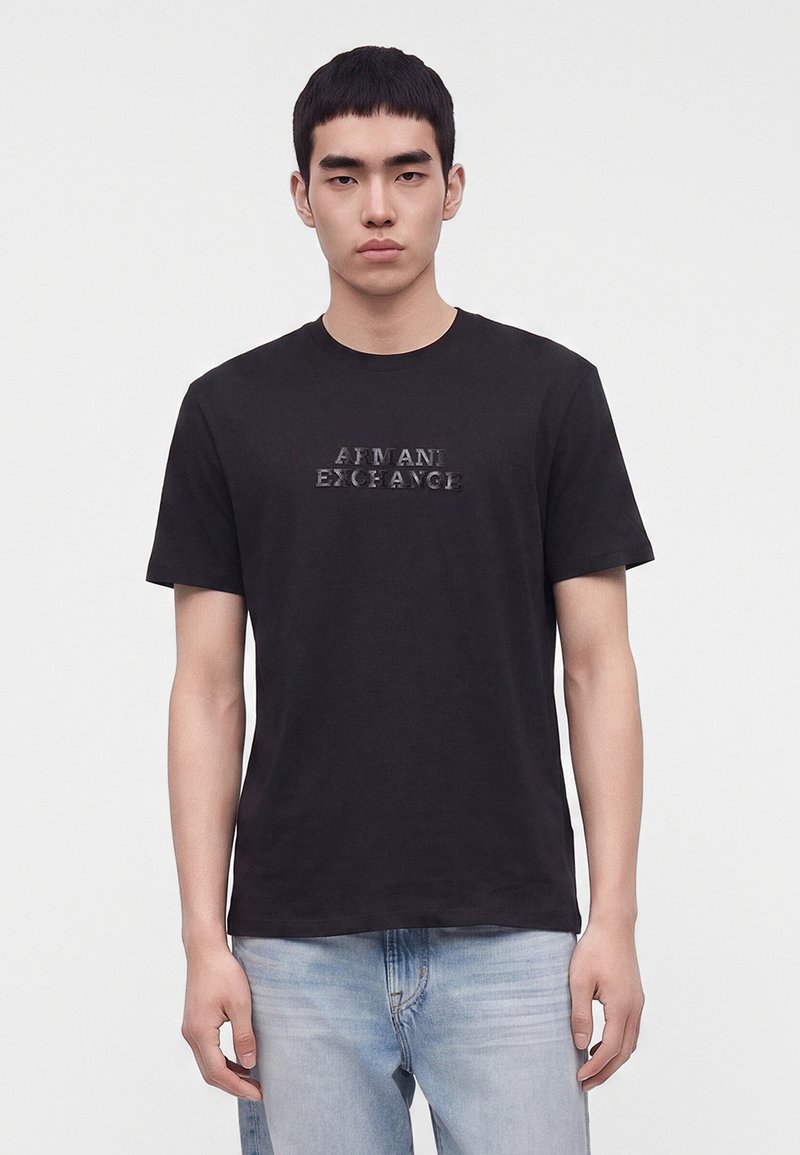 Jeune homme aux cheveux courts noirs portant un t-shirt noir Armani Exchange et un jean bleu clair, se tenant devant un fond clair uni.