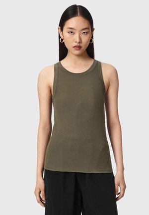 Jonge vrouw met stijl zwart haar, gouden creooloorbellen, een mouwloze olijfgroene geribde top en zwarte broek, staand tegen een grijze achtergrond.