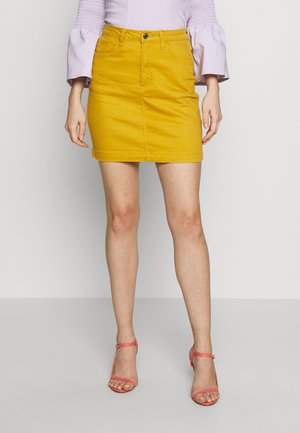 Jupe en denim jaune avec poches avant, une silhouette cintrée et une fermeture à bouton. Assortie à un haut violet clair et des talons roses à lanières.