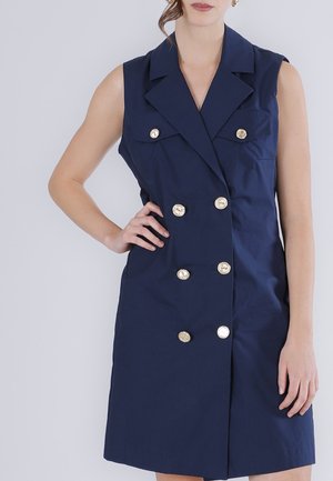 Nainen pukeutuneena hihattomaan, tummansiniseen, kaksiriviseen mekkoon, jossa on kultaiset napit, seisoo kevyttä taustaa vasten, toinen käsi lanteilla.