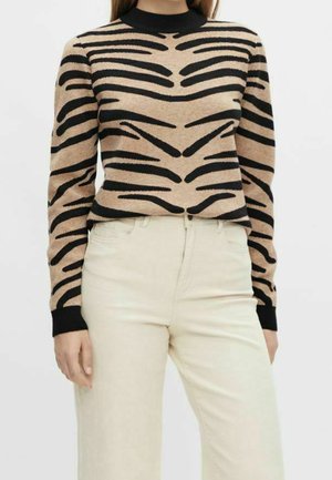 Femme portant un pull à manches longues beige et noir à rayures zèbre, associé à un pantalon beige clair taille haute.