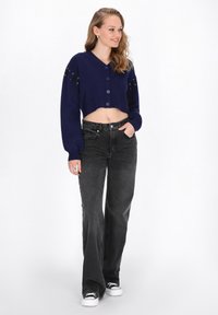 Cardigan cropped blu navy con bottoni e dettagli decorativi; abbinato a jeans neri a vita alta e sneakers nere.