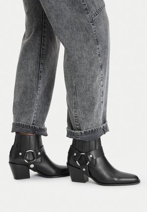 Stivaletti neri alla caviglia con punta a occhiello, realizzati in pelle liscia. Presentano un tacco spesso e dettagli con fibbia in metallo. Indossati con jeans grigi risvoltati.