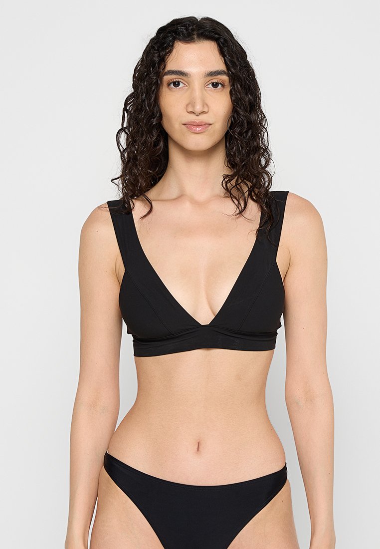 Frau mit lockigem dunklem Haar, die ein schwarzes Bikini-Oberteil mit tiefem V-Ausschnitt und passende Bikini-Hosen trägt, vor einem schlichten weißen Hintergrund.