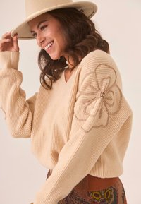 Suéter beige acanalado con escote en V y bordado floral en las mangas, llevado con una falda estampada y un sombrero de fieltro de color claro.