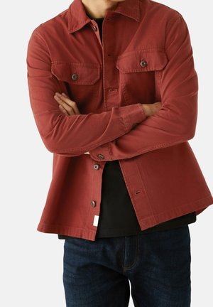 Man met gekruiste armen, gekleed in een roestkleurig overhemdjack, een zwart shirt eronder en donkerblauwe spijkerbroek, tegen een witte achtergrond.