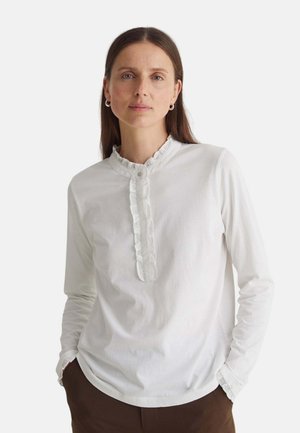 Mujer con cabello largo y castaño vistiendo una camiseta blanca de manga larga con cuello y tapeta con volantes, de pie con las manos en los bolsillos de pantalones marrones.