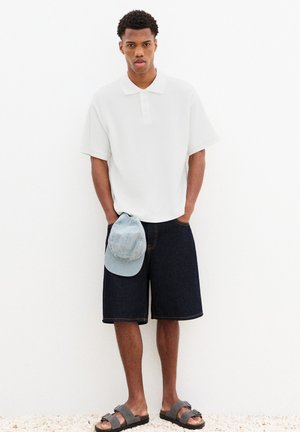 Jeune homme debout les mains dans les poches, portant un polo blanc, un short foncé jusqu'aux genoux, des sandales grises, tenant une casquette bleu clair.