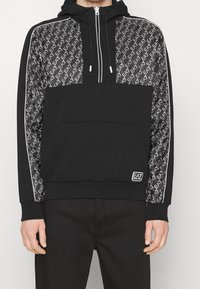 Svart hoodie med dragkedja, med en framficka i kangurostil, prytt med ett mönster av svart och vitt logo på ärmarna, samt vita detaljer längs sidorna.