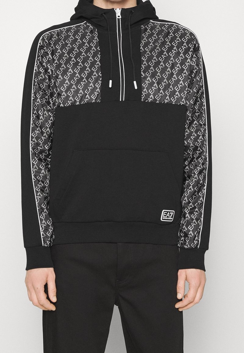 Svart hoodie med dragkedja, med en framficka i kangurostil, prytt med ett mönster av svart och vitt logo på ärmarna, samt vita detaljer längs sidorna.