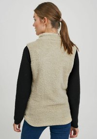 Bežs vilnas veste ar augsto apkakli, teksturētu audumu un melnām garajām piedurknēm. Neformāls dizains, pieguļoša forma un noapaļots apakšmalā aizmugurē.
