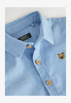 Camisa azul clara de manga larga hecha de tela de mezclilla. Presenta botones marrones y una cara de oso bordada cerca del pecho. Etiqueta visible en el interior del cuello.