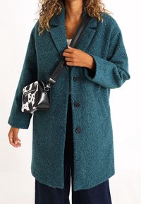 Abrigo texturizado en color azul verdoso con un cuello, botones grandes y bolsillos laterales; combinado con un pequeño bolso cruzado de piel sintética en estampado de vaca en blanco y negro.