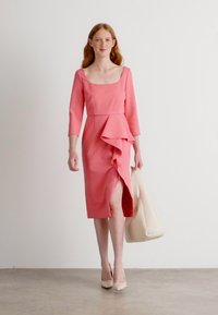 Robe midi rose avec un décolleté carré, des manches trois-quarts, des détails de volants asymétriques et une fente sur le côté, associée à un sac à main crème.
