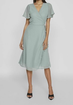 Robe portefeuille verte avec des manches courtes évasées, présentant un motif en chevrons. Fabriquée en tissu léger, longueur au genou, portée avec des talons ouverts noirs.