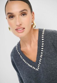 Maglione grigio a V con accentuazioni lucide a forma di diamante attorno al colletto, texture morbida e vestibilità rilassata. Modello che indossa orecchini dorati.