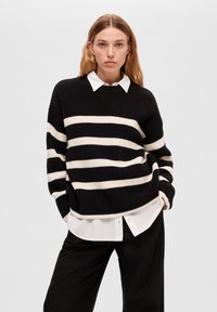 Maglione a righe bianco e nero in maglia con una vestibilità ampia, dotato di un collo rotondo e indossato sopra una camicia bianca con colletto. Pantaloni neri.