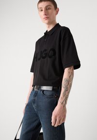 Juodas polo marškinėliai su dideliu matinio paviršiaus "HUGO" logotipu, trumpomis rankovėmis, derinami su tamsiai mėlynomis džinsomis ir pilku diržu.