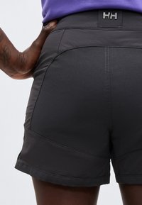 Άτομο που φορά μαύρα σορτς Helly Hansen, με το χέρι ακουμπισμένο στο ισχίο, εμφανιζόμενο από τη μέση πίσω μέχρι το μέσο του μηρού.