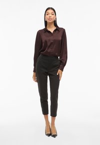 Satenska burgundska bluza z gumbastim zapiranjem, dolgimi rokavi in črnimi kratkimi hlačami, v kombinaciji s črnimi čevlji na visoki peti z zlatim detajlom.