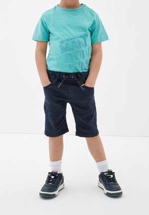 T-shirt en coton turquoise avec texte en relief, associé à un short bleu marine avec taille réglable et poches latérales. Baskets noires avec chaussettes blanches.