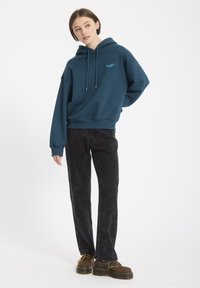 Turkusowy bawełniany hoodie ze sznurkami, z małym niebieskim logo. W zestawie z czarnymi spodniami z sztruksu i brązowymi butami na sznurowadła.