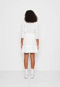 Mujer con cabello rizado de espaldas, vistiendo un vestido blanco con volantes, calcetines blancos y zapatillas blancas, contra un fondo simple.