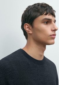 Junger Mann mit kurzen dunklen Haaren, der einen dunkel strukturierten Pullover trägt und seitlich vor einem schlichten hellen Hintergrund schaut.