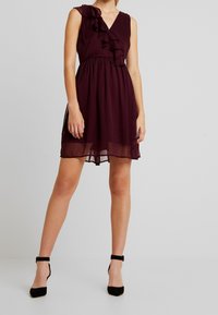 Burgundröd ärmlös klänning med en volang på vänster axel, V-ringning och en samlad midja, tillverkad i lätt tyg. Svarta högklackade skor.
