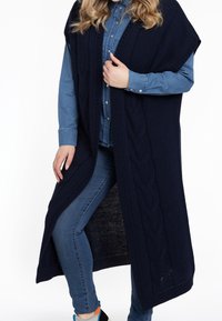 Długa granatowa dzianinowa kamizelka z wzorem warkocza, otwartym przodem i rozcięciami po bokach, noszona na koszuli jeansowej i niebieskich dżinsach.