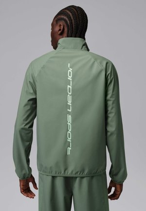 Mężczyzna z warkoczami, ubrany w jasnolistopadową kurtkę JORDAN SPORT i pasujące spodnie, stoi tyłem na szarym tle.