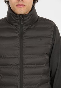 Svart pufferväst med hög krage, framzipper och matt finish. Har horisontell sömnad och en liten logotyp på bröstet.