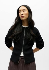 Veste noire à fermeture éclair avec une coupe structurée, un col rond et des manches trois-quarts, portée avec un haut clair et une jupe marron.