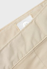 Beige stof med en glat overflade, der viser en nærbillede af en knap og et mærke. Inkluderer synlig syning og en sømdetalje.