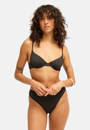 Femme aux cheveux bouclés portant un bikini noir avec un bas taille haute, posant les mains jointes à la taille, sur un fond clair uni.