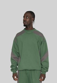 Ein schwarzer Mann mit geflochtenem Haar, der einen übergroßen dunkelgrünen und grauen Sweatshirt sowie passende Hose trägt, schaut nach links vor einem schlichten grauen Hintergrund.