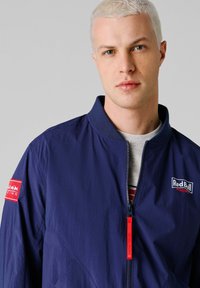 Jonge man met kort blond haar, gekleed in een marineblauw Red Bull Racing-jack, dichtgeritst over een lichtgrijs shirt, neutrale achtergrond.