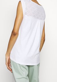 Top blanco sin mangas con un diseño decorativo de ojales en la parte del cuello, de corte holgado y tela suave. Combinado con pantalones verde claro.