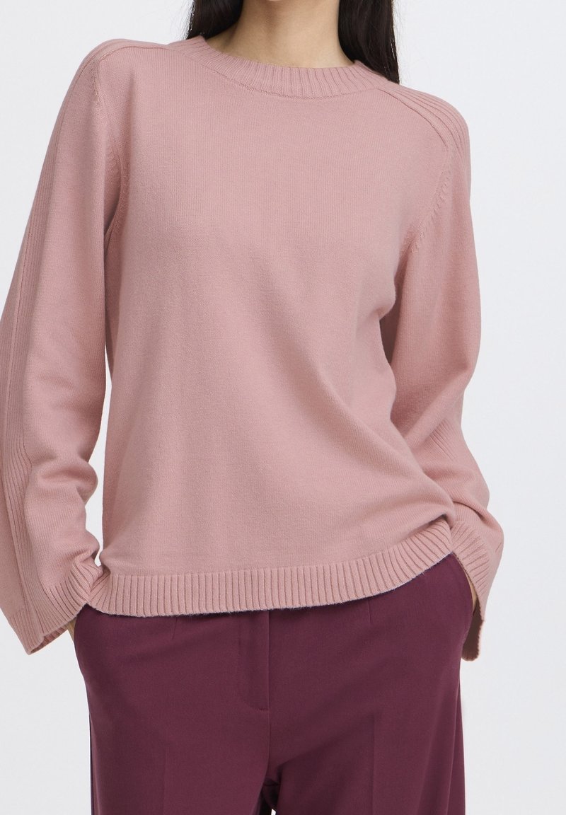 Femme portant un pull en maille rose clair avec des bords côtelés et des manches longues, associé à un pantalon violet foncé, les mains dans les poches.