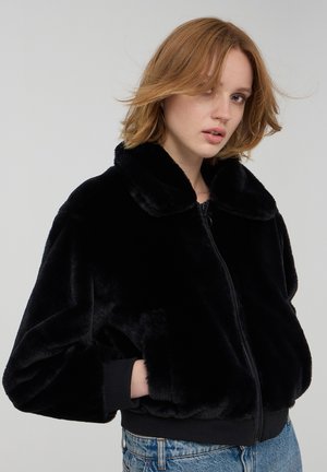Czarna kurtka bomber z sztucznego futra z zapięciem na zamek, okrągłym kołnierzem i ściągaczami. Miękka tekstura, luźny krój i kieszenie boczne.