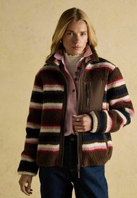 Fleecejacke mit horizontalen Streifen in Braun, Weiß, Pink und Blau, mit einem Reißverschluss und einer Brusttasche. Über einem Hemd mit Kragen getragen.